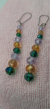 Load image into Gallery viewer, A pair of dangling beaded earrings with faceted beads in green, amber, lavender, and iridescent blue, hanging from silver fishhook wires on a textured pink fabric background.

